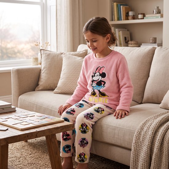 Meisje zittend op een bank in een roze koraalfleece pyjama met Minnie Mouse opdruk, tekst MINNIE en een Minnie-broek.