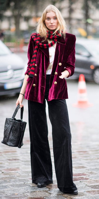 Model draagt een wijnrode fluwelen blazer met dubbele rij gouden knopen, zwarte wijde broek en rood-zwarte gestreepte sjaal.