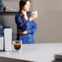 Espresso in een glazen kopje naast een De'Longhi waterfilter, met een persoon op de achtergrond.