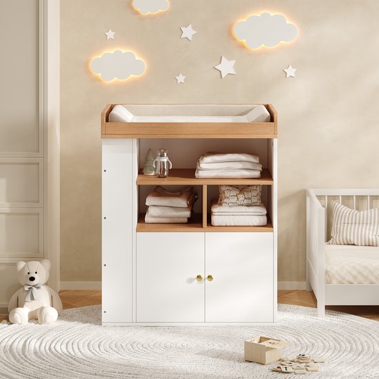 Witte commode met houten accenten en aankleedkussen, gevuld met gevouwen babykleding en items in een kinderkamer.