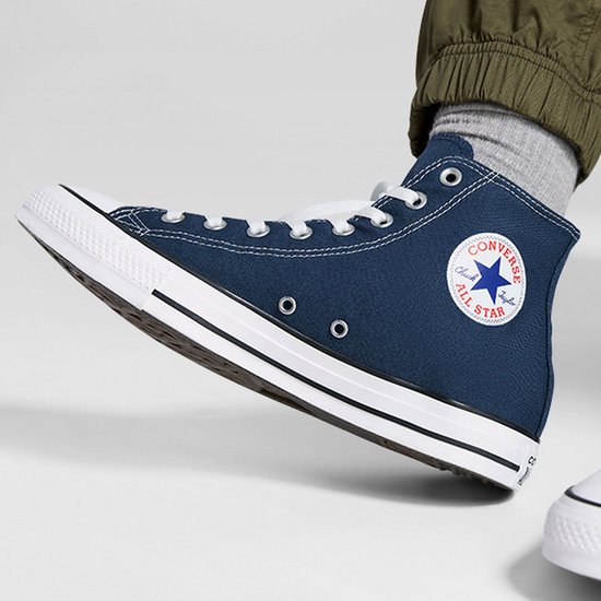Voet met donkerblauwe Converse All Star sneaker, van de zijkant en iets van voren gezien. Met grijze sok en groene broek.