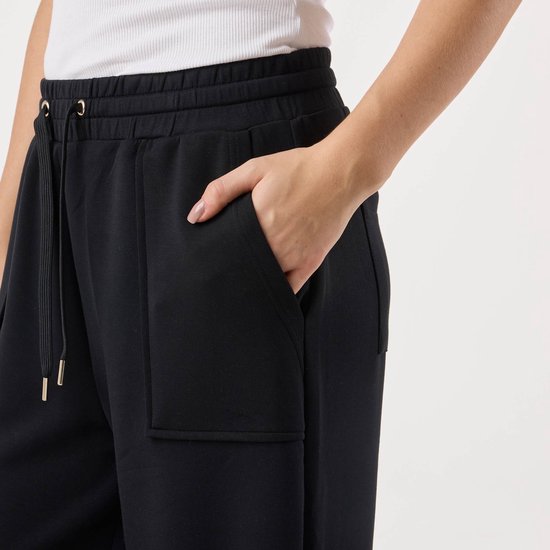 Close-up van een zwarte broek met hoge taille, koord met gouden uiteinden en opgestikte zakken.
