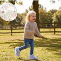 Lachend kind in beige trui en jeans, houdt een doorzichtige ballon met gekleurde confetti vast in een park.