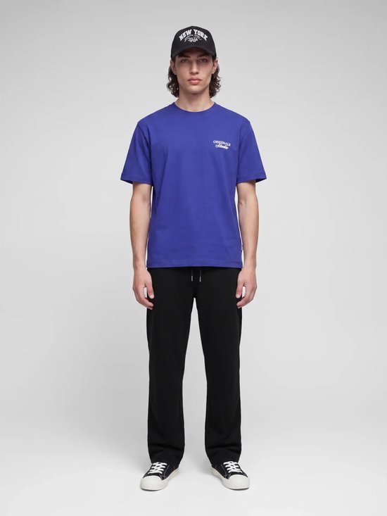 Model staat rechtop in een blauw T-shirt met 'ORIGINALS Studio' opdruk, zwarte pet en zwarte joggingbroek.