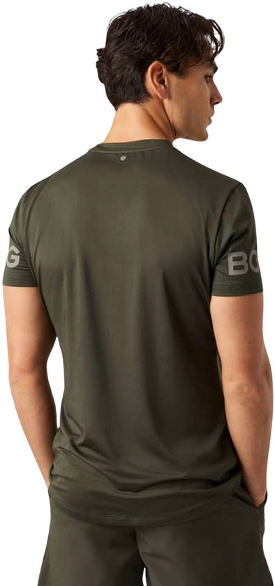 Donkergroen sportshirt van gerecycled polyester, met logo op de mouw. Man met handen in zakken.
