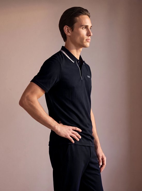 Man draagt een donkerblauw poloshirt met verticale ribbels, geborduurd logo op de borst, en bijpassende broek.