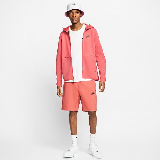 Model draagt koraalroze Nike Tech Fleece-hoodie en short met witte hoed en sneakers.