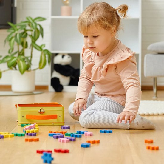 Een jong kind met een roze trui speelt met Plus-Plus blokken op een houten vloer.