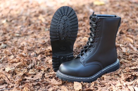 Bottes de combat noires en cuir avec coutures blanches et lacets. Une botte est debout, l'autre est couchée avec la semelle vers le haut. Entourées de feuilles brunes.