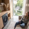 Lave-linge gris dans un salon lumineux, à côté d'une femme écoutant de la musique avec un casque sur le canapé.