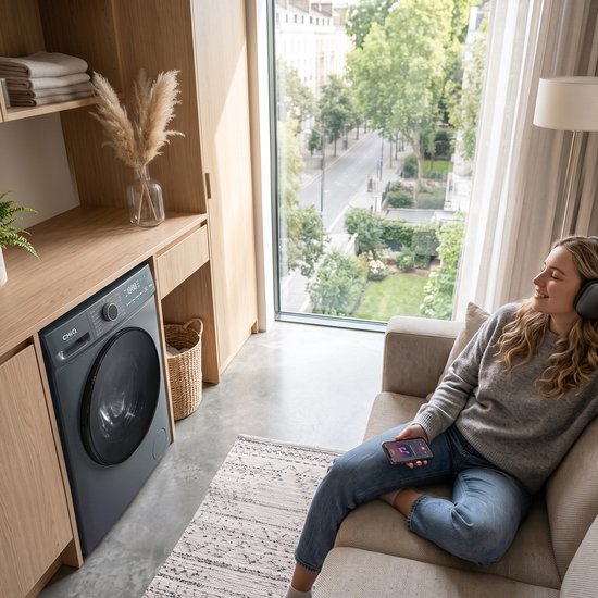 Lave-linge gris dans un salon lumineux, à côté d'une femme écoutant de la musique avec un casque sur le canapé.