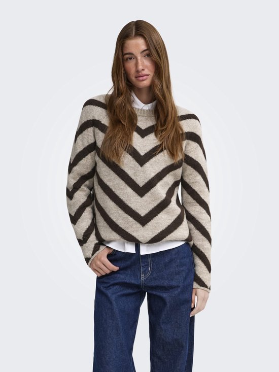 Model draagt een beige gebreide trui met donkerbruin chevronpatroon over een wit overhemd en blauwe jeans.