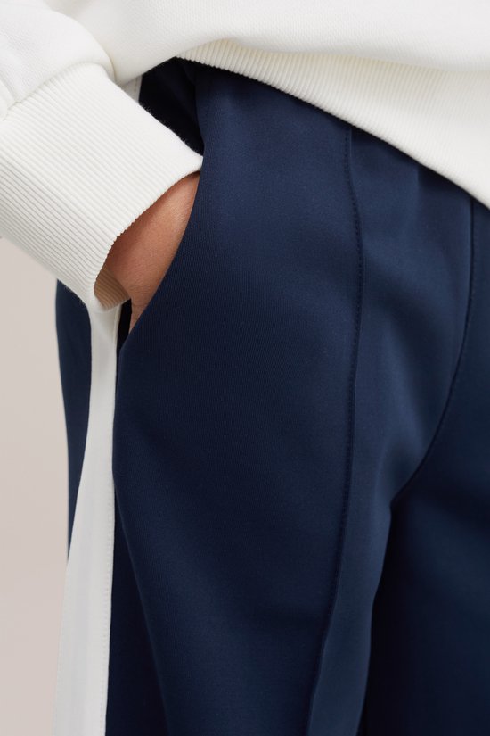 Detailopname van een donkerblauwe broek met witte bies, gedragen door een model in een witte trui.