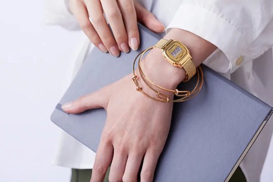 Goudkleurig Casio digitaal horloge met mesh-band om pols met gouden armbanden, houdend een grijs notitieboek.