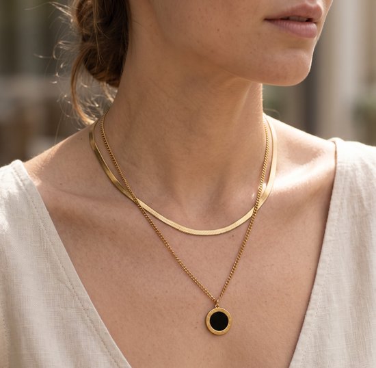 Model draagt een gelaagde gouden ketting met een platte slangenketting en een hanger met zwarte ronde steen.