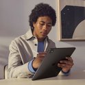 Een persoon met donker krullend haar gebruikt een stylus op een donkergrijze tablet aan een bureau.
