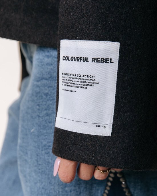 Detail van een label op de mouw van een donkerbruine wollen jas met de tekst 'COLOURFUL REBEL WOMENSWEAR COLLECTION'.