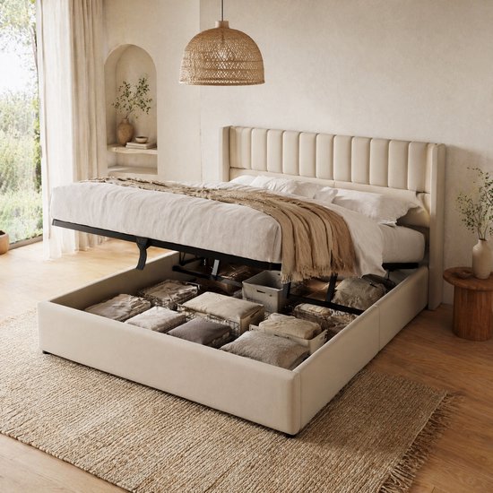 Een beige gestoffeerd bed met omhoog geklikt matras, wat extra opbergruimte met hydraulische lift toont. Witte lakens en dekens zijn zichtbaar.