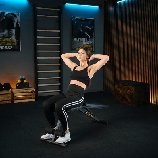 Vrouw met zwarte sportkleding doet een sit-up op een zwarte fitnessbank in een sportschool.