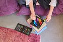 Persoon zit op een paarse poef met het IQ Circle spel op boeken. Ze legt gekleurde stukken in een cirkel. Twee uitdagingenboeken liggen open.