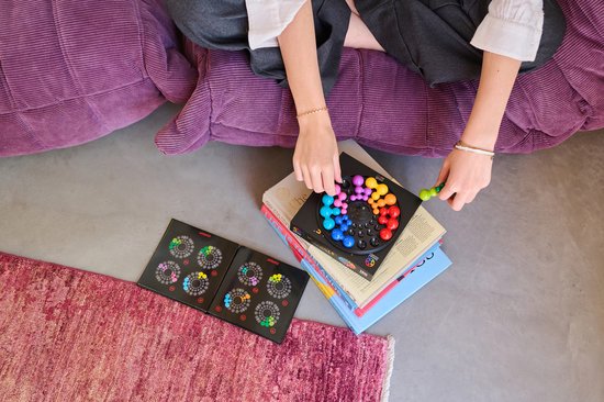Persoon zit op een paarse poef met het IQ Circle spel op boeken. Ze legt gekleurde stukken in een cirkel. Twee uitdagingenboeken liggen open.