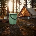 Groene campingtoilet emmer met deksel, naast een tent en een kampvuur in een bos.