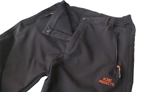Bovenzijde van een zwarte softshell broek met open rits en oranje 'ACME PROJECTS' logo.