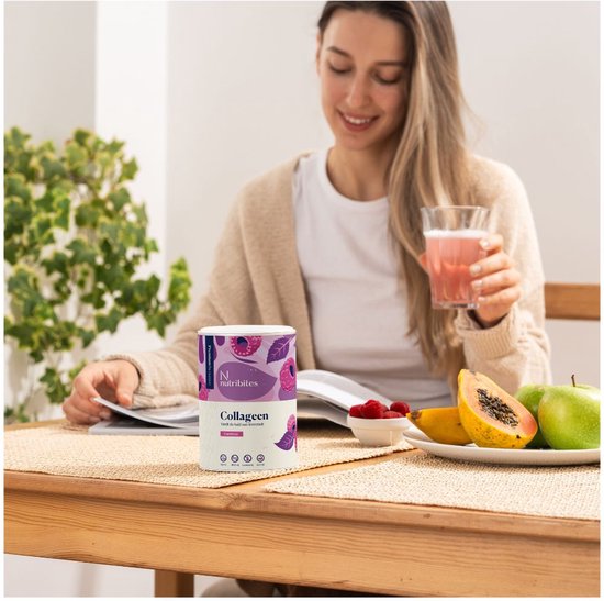 Vrouw leest en drinkt aan tafel, naast een blik Nutribites collageenpoeder framboos en vers fruit. Een glas met roze drankje in haar hand.