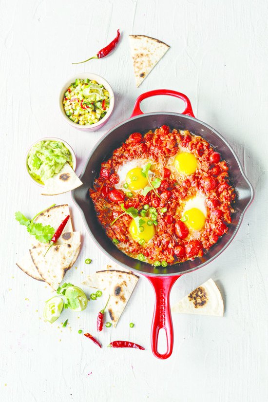 Kersrode koekenpan gevuld met gebakken eieren in tomatensaus, met guacamole, salsa, tortilla's, limoen en chilipepers ernaast.