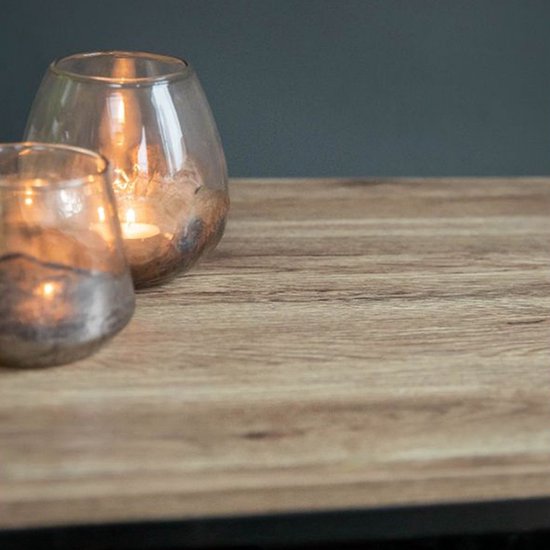 Close-up van het licht houten tafelblad met een grof patroon. Twee glazen windlichten met brandende kaarsen erop.