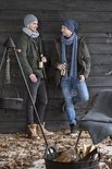 Twee mannen met mutsen en sjaals, staand tegen een houten muur, met flessen bier in hun handen.