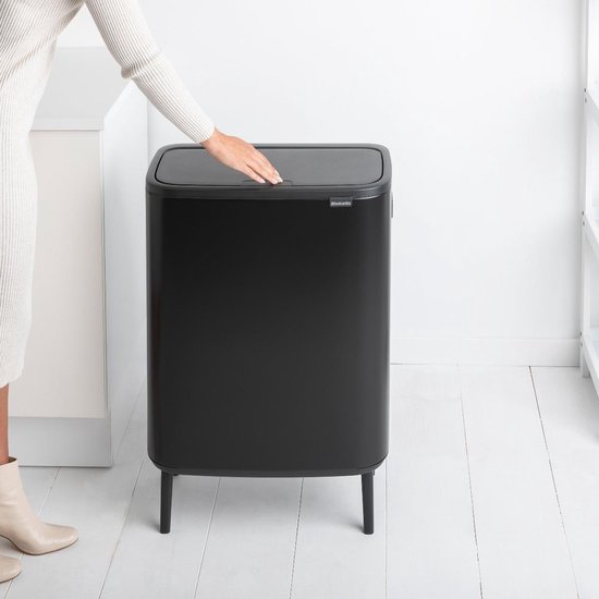 Une femme en robe blanche tricotée et bottines beiges touche le couvercle fermé d'une poubelle Brabantia noire.