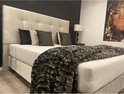 Tweepersoons boxspring in beige velours met een gestoffeerd hoofdbord, vier kussens en een donkergrijze sprei.