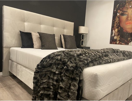 Tweepersoons boxspring in beige velours met een gestoffeerd hoofdbord, vier kussens en een donkergrijze sprei.