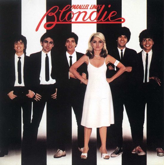 Bandleden van Blondie in pakken, zangeres Debbie Harry in een witte jurk, tegen een zwart-wit gestreepte achtergrond.