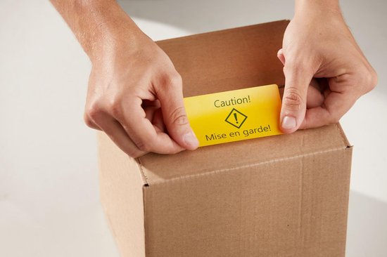 Twee handen plakken een gele waarschuwingssticker "Caution! Mise en garde!" op een kartonnen doos.