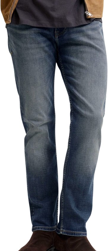 Man in spijkerbroek, bruine jas en donker T-shirt, zicht op de voorkant.