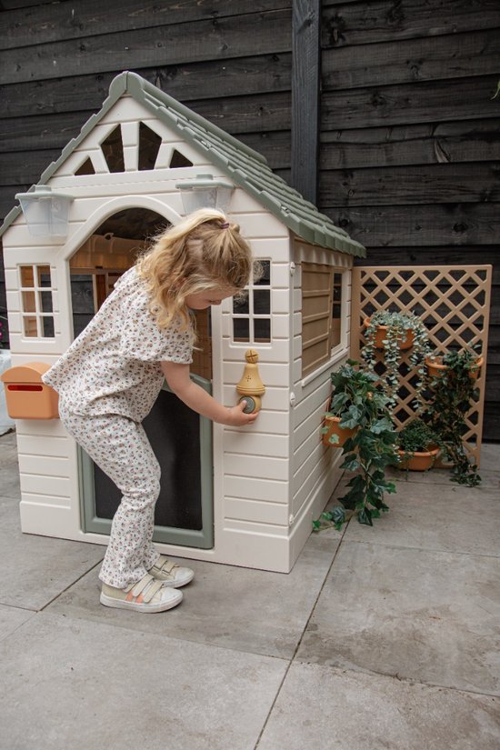 Meisje met blond haar in een speelhuisje met groen dak, een brievenbus en planten ernaast.