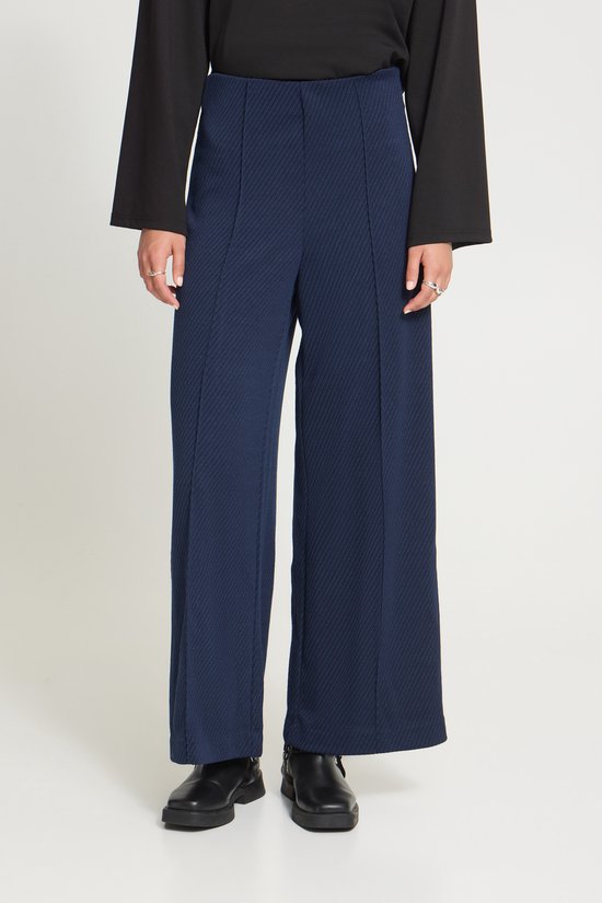 Model draagt een donkerblauwe broek met wijde pijpen, diagonaal patroon en zwarte schoenen.