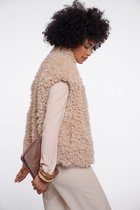 Model draagt een beige, pluizige teddy gilet over een lichtbruine coltrui. Ze houdt een bronzen clutch en gouden armbanden vast.