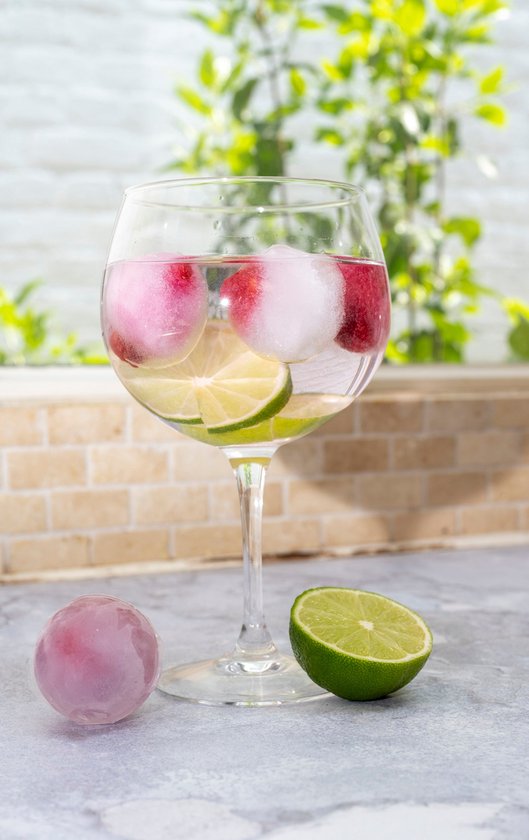 Een glas met drank, grote ijsbollen, partjes limoen en een roze ijsbol.