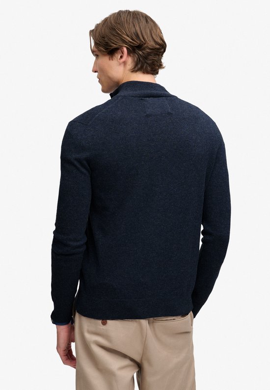Achterkant van een man met donkerblauwe melange trui met ritssluiting, lichtblauw T-shirt en beige broek.