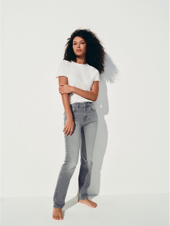 Model in een witte t-shirt en grijze jeans, staand tegen een witte muur.