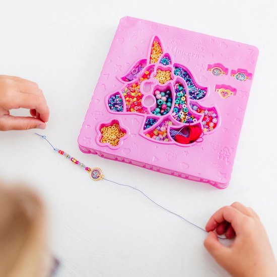 Kinderhanden maken een armband met kralen en tassels. Een roze knutseldoos met eenhoornvormige vakjes vol kralen.