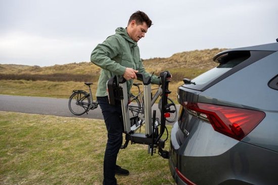 Man in lichtgroene jas bevestigt een zilverkleurige trekhaakfietsendrager aan de achterkant van een auto.