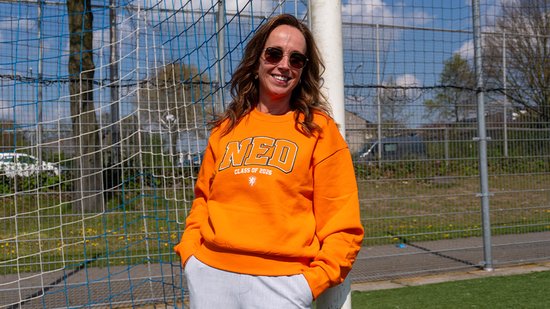 Vrouw met zonnebril draagt een oranje trui met "NED CLASS OF 2026" opdruk.