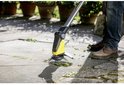 Een man met spijkerbroek en bruine schoenen gebruikt de Kärcher onkruidverwijderaar op een betegeld terras.