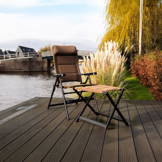 Bruine, verstelbare campingstoel en voetenbankje met donker metalen frame, op een houten terras aan het water.