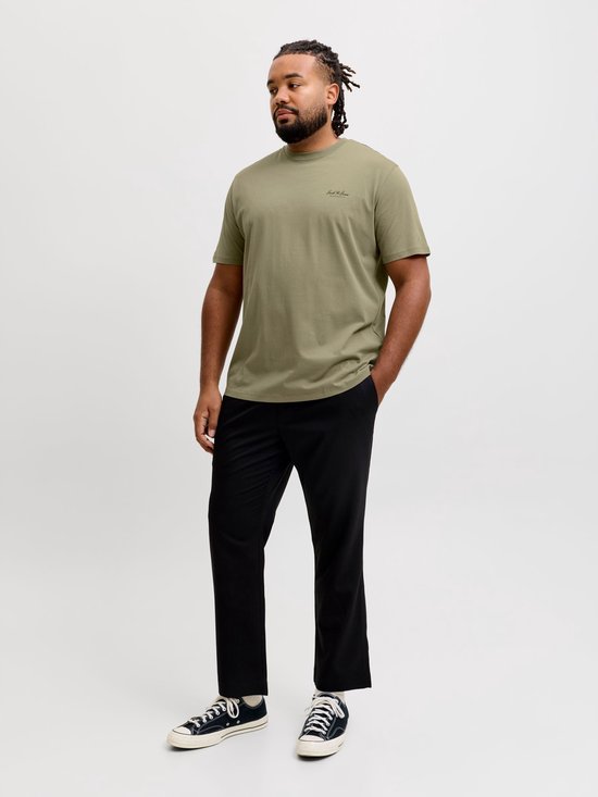 Man draagt olijfgroen T-shirt met 'Jack & Jones ARCHIVE SELECTION' logo en zwarte broek. Hand in zak.
