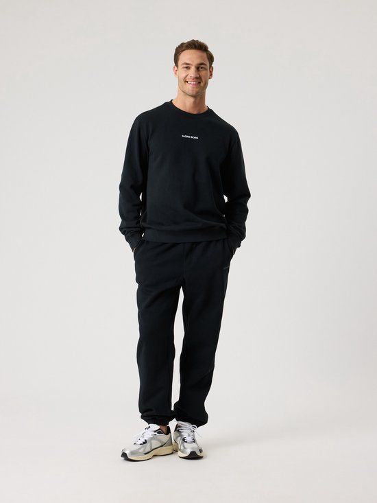 Man in een zwarte sweater en bijpassende broek, lachend met handen in de zakken.